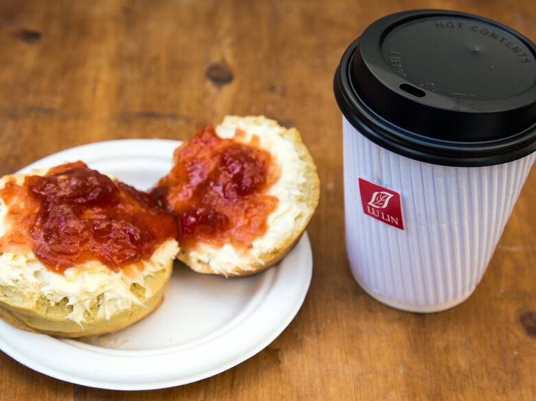 Photo of a scone covered in clotted cream on the bottom and jam on top, next to a takeaway cup of tea, served by Tea, Bread & Brownie at Southbank Centre Food Market