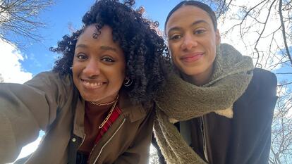 Two black women lean down to face camera, smiling; blue sky in background