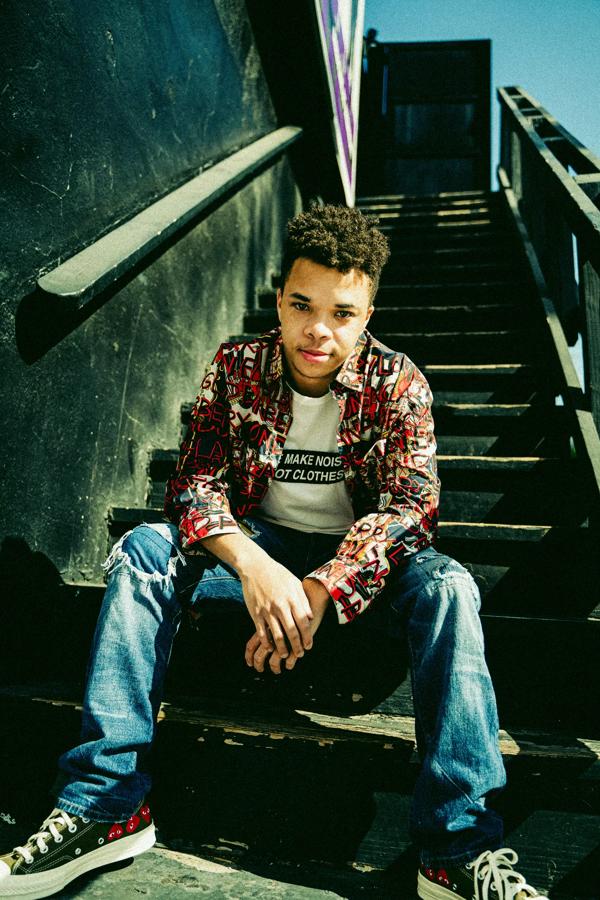 Musician amorphous sits on a staircase outside wearing a patterned shirt, white slogan Tshirt, blue jeans and a pair of converses.