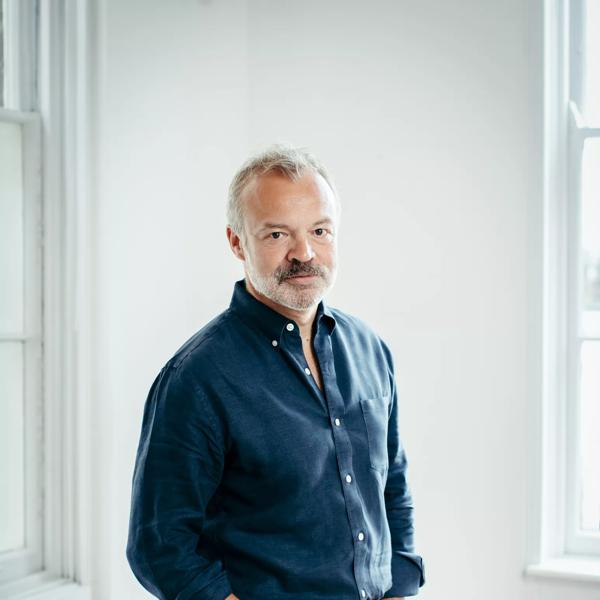 Graham Norton, author and talkshow host, standing in a white room wearing dark blue clothing