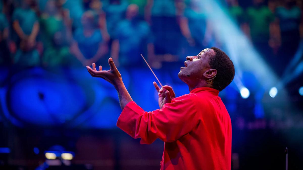 Head and shoulders shot of conductor Kwamé Ryan on stage, mid performance. He wears a red shirt, a baton in his right hand