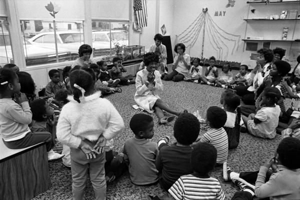Congresswoman Shirley Chisholm, a Black woman with her hair in a beehive hairstyle, sits on the floor as she plays with children at South Central Community Child Care Center, Los Angeles in 1972