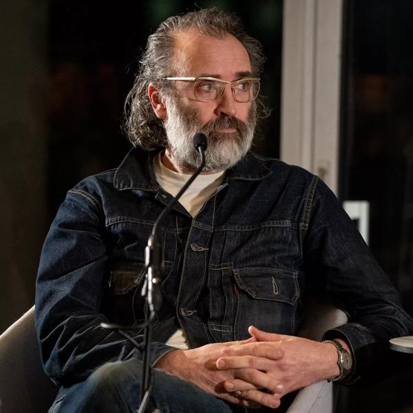 Mike Nelson seated behind a microphone; he has a grey beard and wears glasses and a dark denim shirt