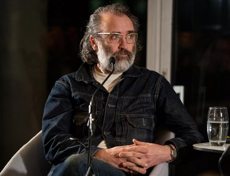Mike Nelson seated behind a microphone; he has a grey beard and wears glasses and a dark denim shirt