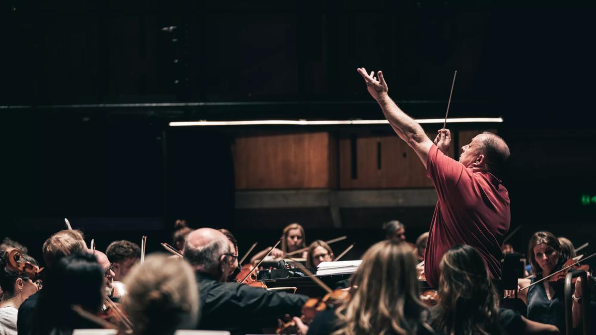 A man in a red shirt stands centre stage and conducts orchestra 