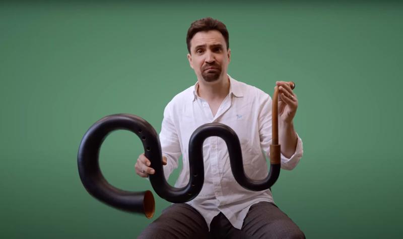 Andrew Kershaw, a white man with dark hair and a goatee beard, of the Orchestra of the Age of Enlightenment sits holding a musical instrument called the serpent, which is a long bendy brass instrument made of wood wrapped in dark leather