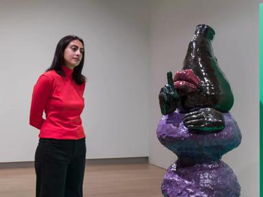 Woman looking at a ceramic figurative sculpture with a purple body and black head, with fingers raised to lips in a shushing gesture. Installation view of Woody de Othello, Strange Clay: Ceramics in Contemporary Art at the Hayward Gallery.