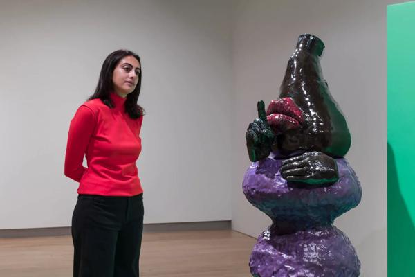 Woman looking at a ceramic figurative sculpture with a purple body and black head, with fingers raised to lips in a shushing gesture. Installation view of Woody de Othello, Strange Clay: Ceramics in Contemporary Art at the Hayward Gallery.