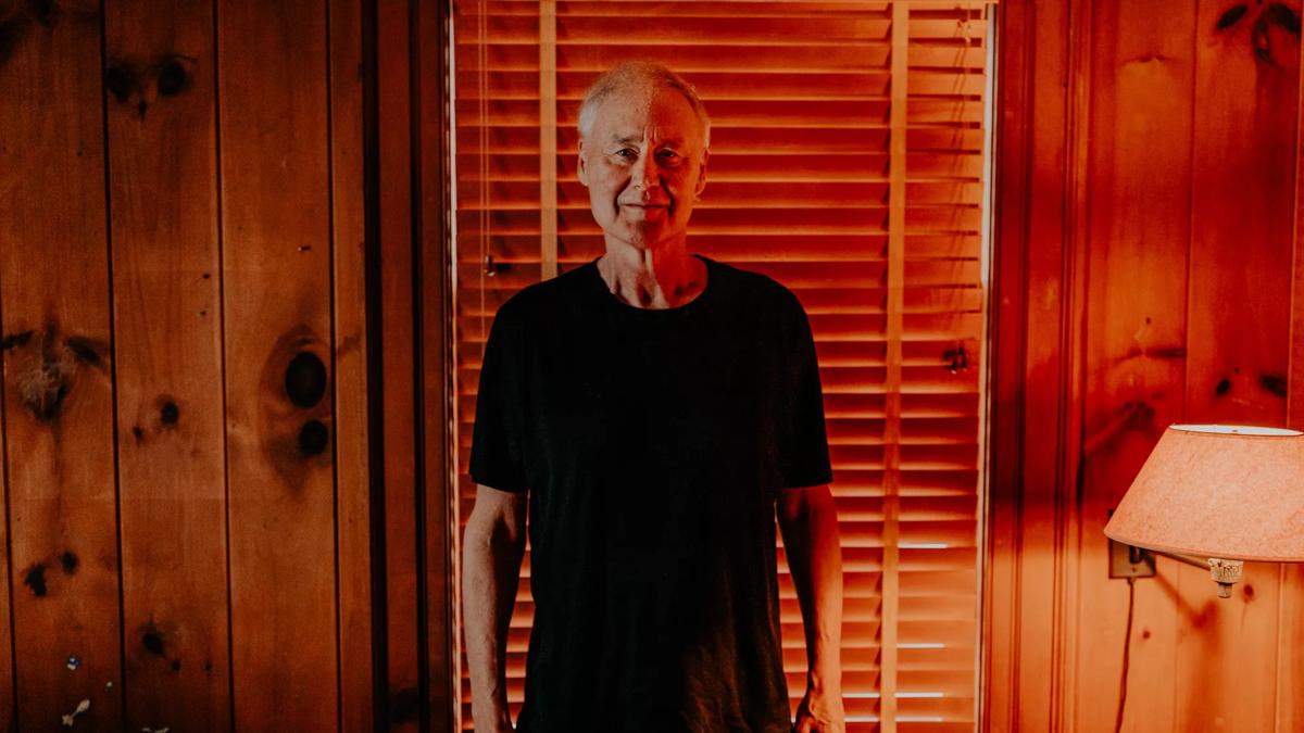 Musician Bruce Hornsby stands in front of a wooden wall with a window covered by blinds, wearing a black T-Shirt.