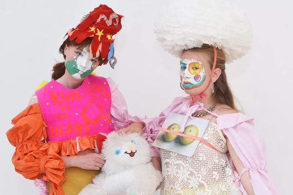 Two young white women with painted faces stand in flamboyant costumes and hold between them a cuddly toy sheep.