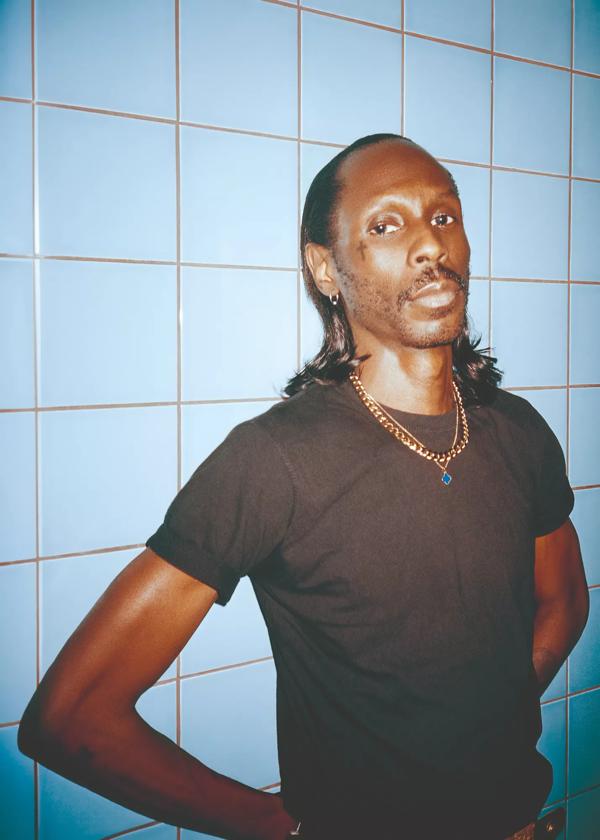 Josh Caffe leans against a blue tiled wall with hands on hips, wearing a black T-shirt and necklace.