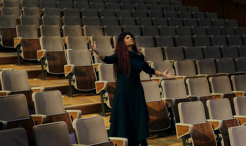 The musician Chaka Khan, a Black woman with long curly hair wears a long black dress and stands with her arms stretched outwards among the seats of the Royal Festival Hall auditorium
