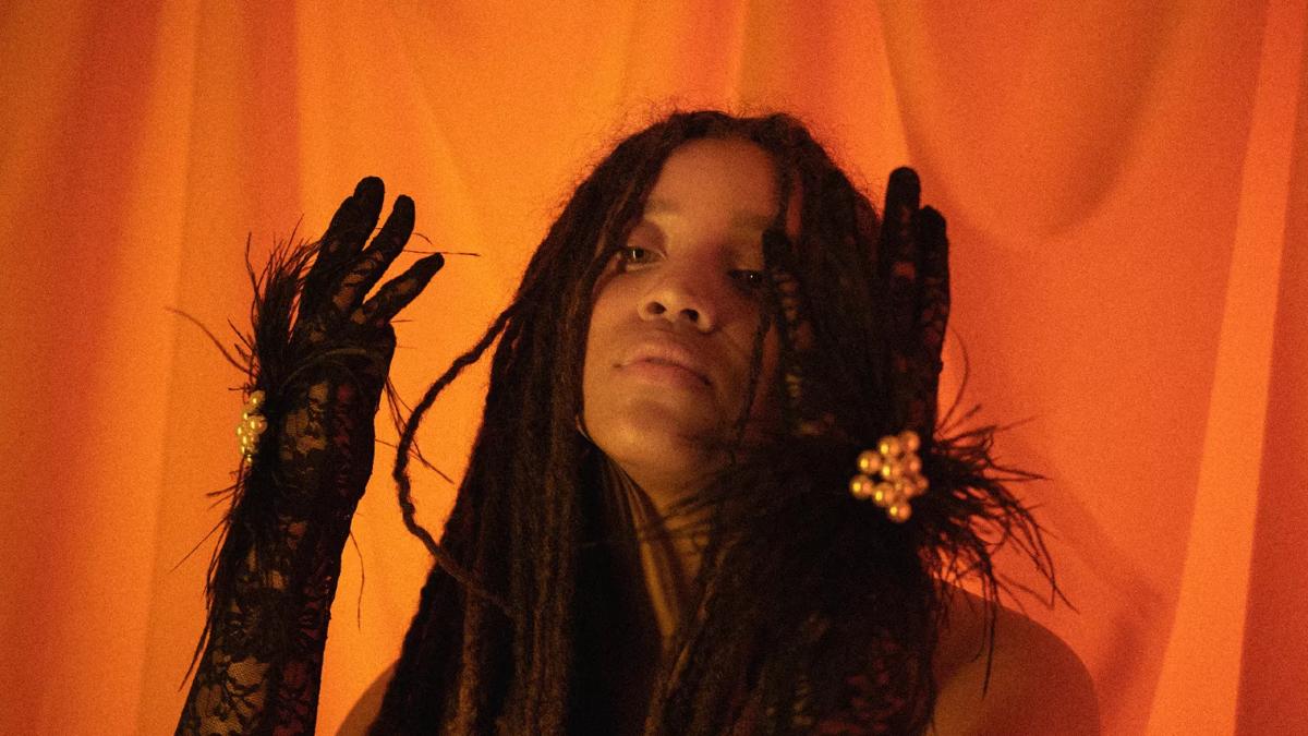 Portrait of a Black woman with her hands raised in front of an orange fabric background