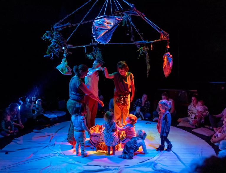 Performers and children stand in a circle with a bright box in the middle