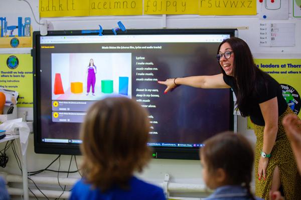 Picture of a teacher pointing to a screen and smiling, in front of young pupils. 