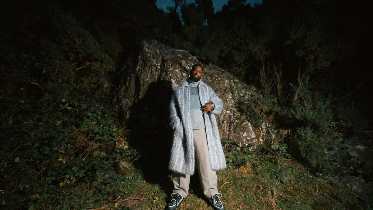 Ghetts stands against a rocky outcrop wearing a light blue jacket. 
