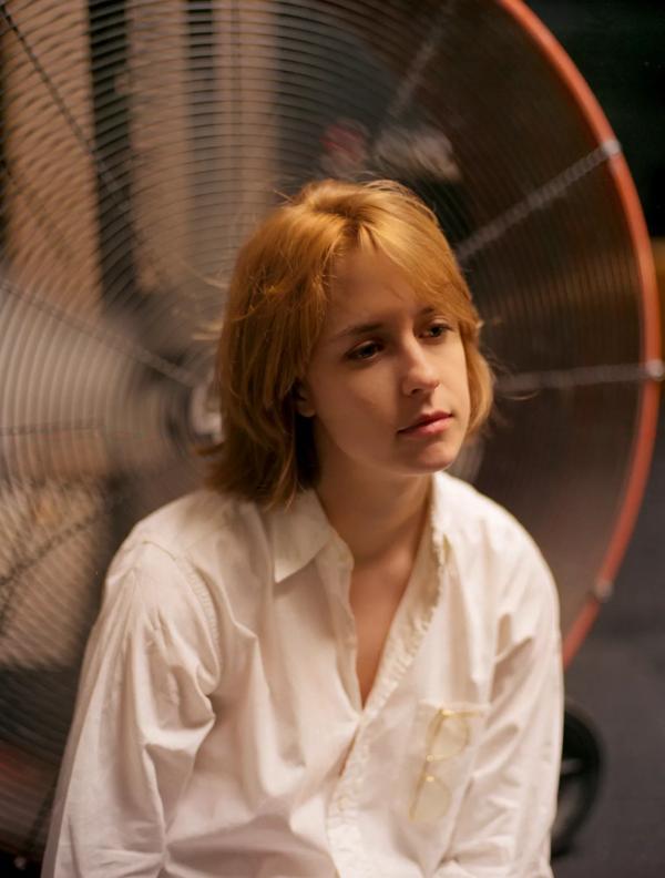Person with ginger hair, wearing a white blouse, posing in front of a fan