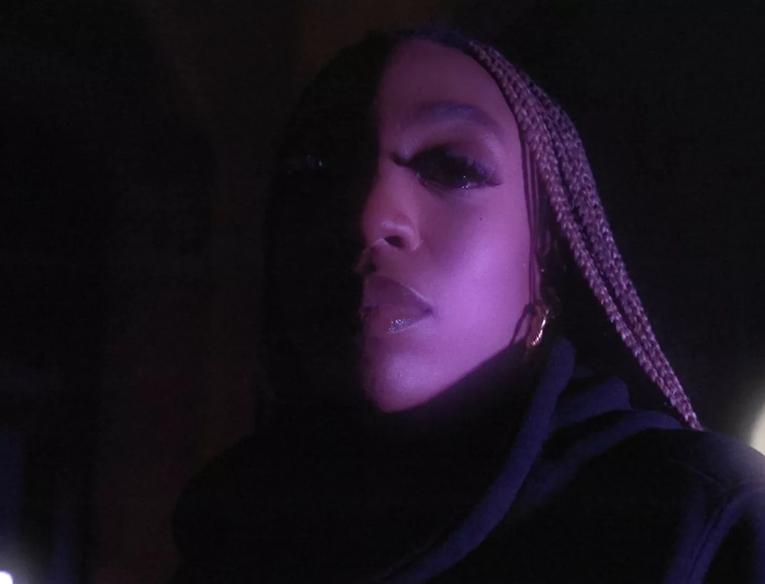 A screenshot photo of Lowlah, a young Black woman with brown skin, long brown braids wearing a black hoodie. Lowlah looks directly into the camera and is lit on one side of her face with purple lighting.