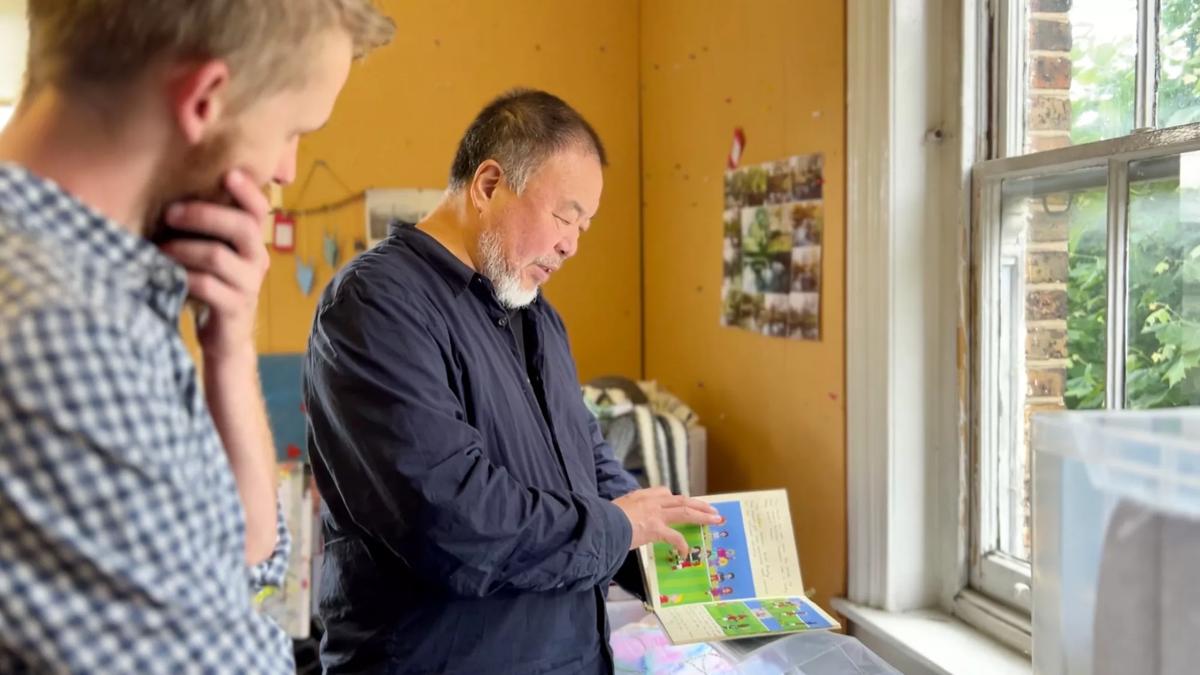 Ai Weiwei stood next to a window looking through a book . The person next to him has his hand over his mouth while gazing at the open book