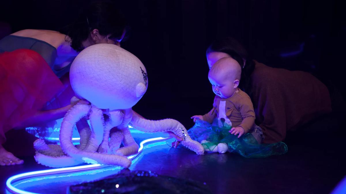 A performer holds an octopus made out of a pink material to a sitting baby  looking at the octopus curiously. LED blue light is on the floor.