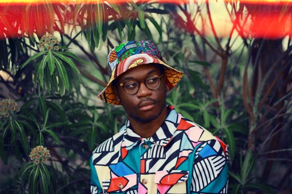 A person wearing a boldly patterned shirt and bucket hat looking to the right with plants behind 