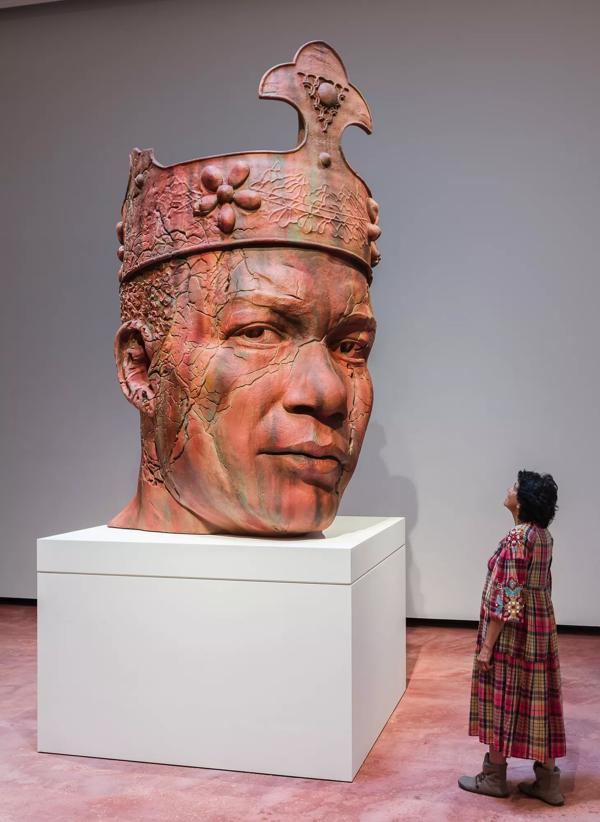 An installation view of a large clay sculpture of a human head wearing a crown and looking to the right.