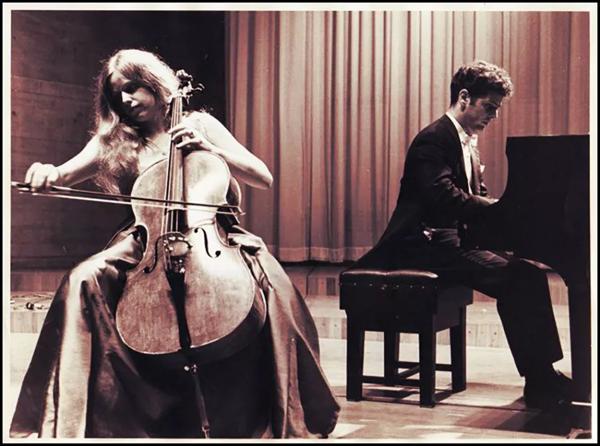 Jacqueline du Pré and Daniel Barenboim perform on stage; du Pré in the foreground seated behind her cello, Barenboim behind her seated at the piano