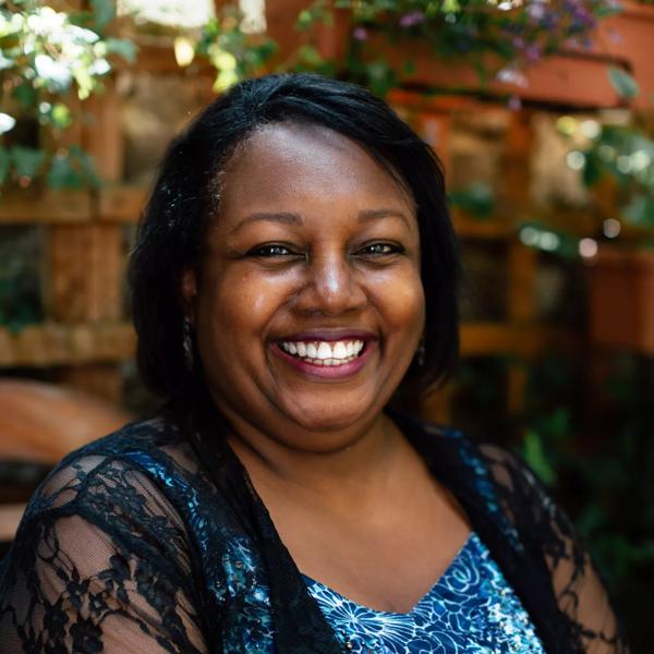 Portrait of author Malorie Blackman. She sits in a garden wearing a patterned blue top and a lace cover up. 