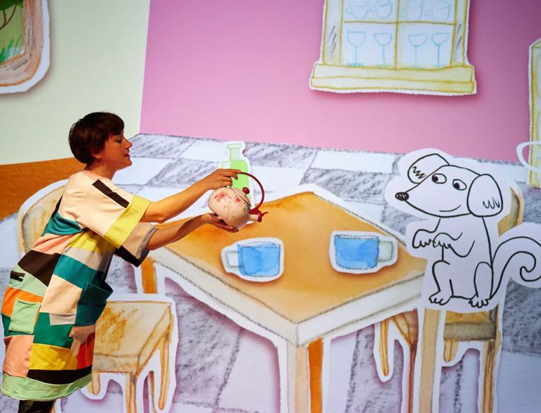 A person pours a teapot in front of a cartoon table setting.