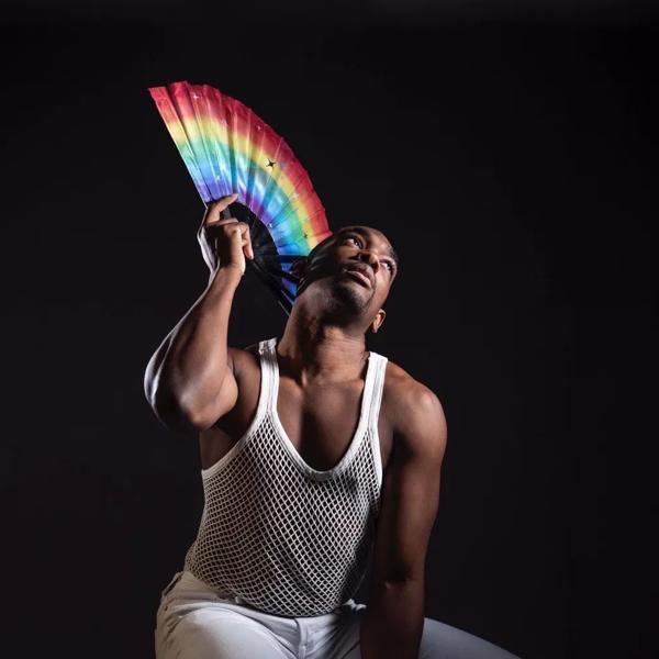 Jay Jay Revlon crouched down with white vest on, holding rainbow coloured fan next to face, eyes looking up, black background