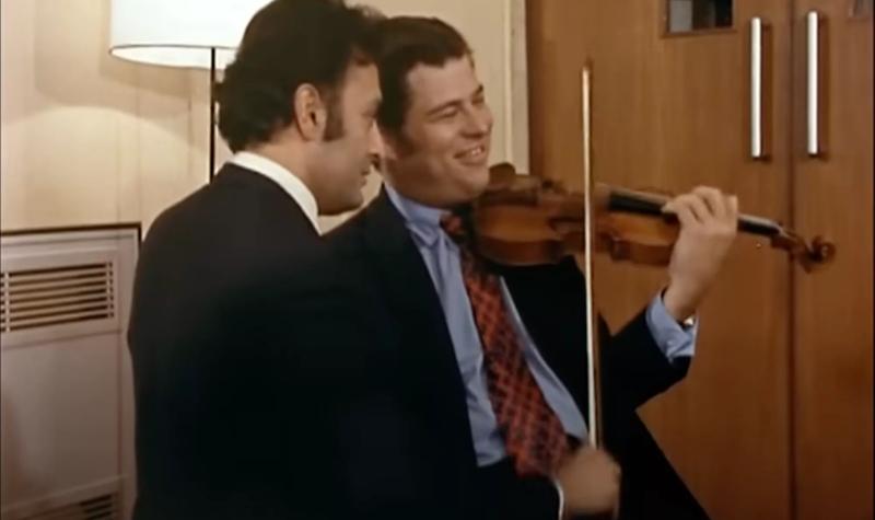 A screengrab from Christopher Nupen's film 'The Trout' that shows Daniel Barenboim & Itzhak Perlman backstage in the Queen Elizabeth Hall, 1969; the two men stand close together whilst playing a single violin; Perlman holds the instrument, and Barenboim the bow.