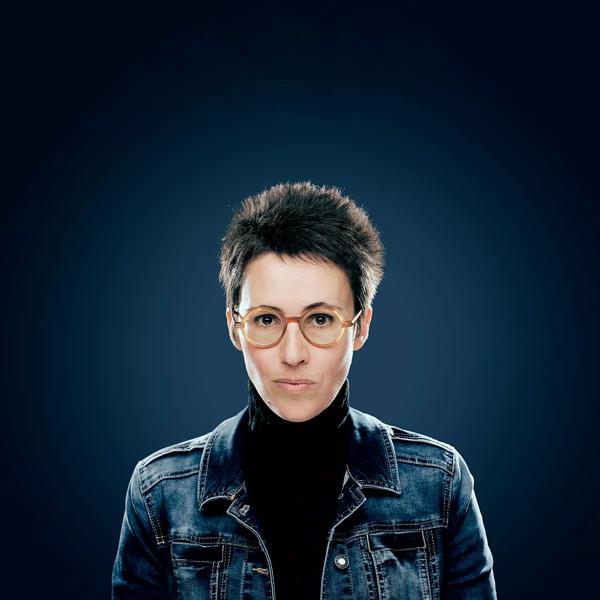 Eva Baltasar, a woman with cropped hair and red glasses, wears a denim jacket and stands against a dark blue backdrop