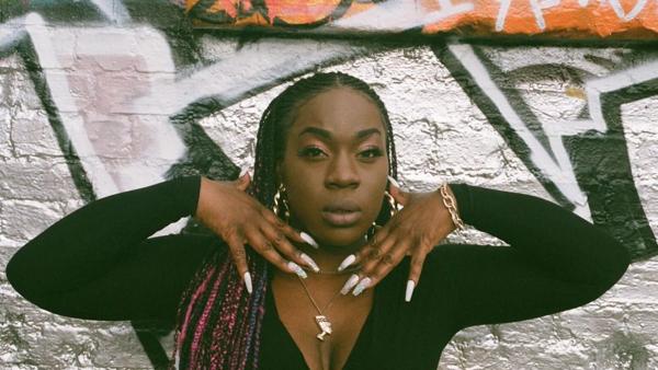 Portrait of a Black woman posing against a grafittied wall with her hands at her face 