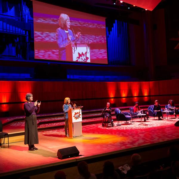 On the left is the BSL interpreter, Jude Kelly in the centre standing behind a podium and on the right Liz Carr in her wheelchair and panellist sitting on armchairs at WOW - Women of the World festival. 