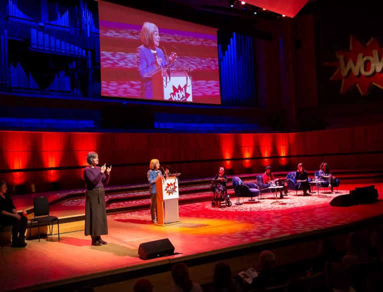 On the left is the BSL interpreter, Jude Kelly in the centre standing behind a podium and on the right Liz Carr in her wheelchair and panellist sitting on armchairs at WOW - Women of the World festival. 