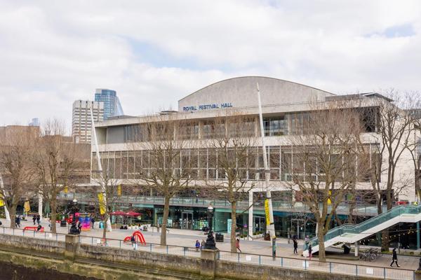 The Royal Festival Hall building viewed from Hungerford Bridge, the trees on the riverside are leafless and some people can be seen walking on Queen's Walk