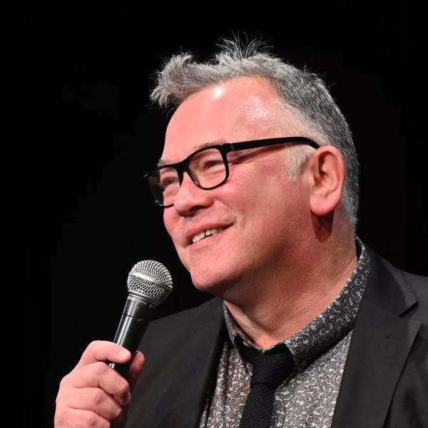A side on profile of comedian Stewart Lee; a grey haired man with thick, black glasses smiles in front of a microphone. He's wearing a black blazer over a grey checked shirt and a slim black tie.  
