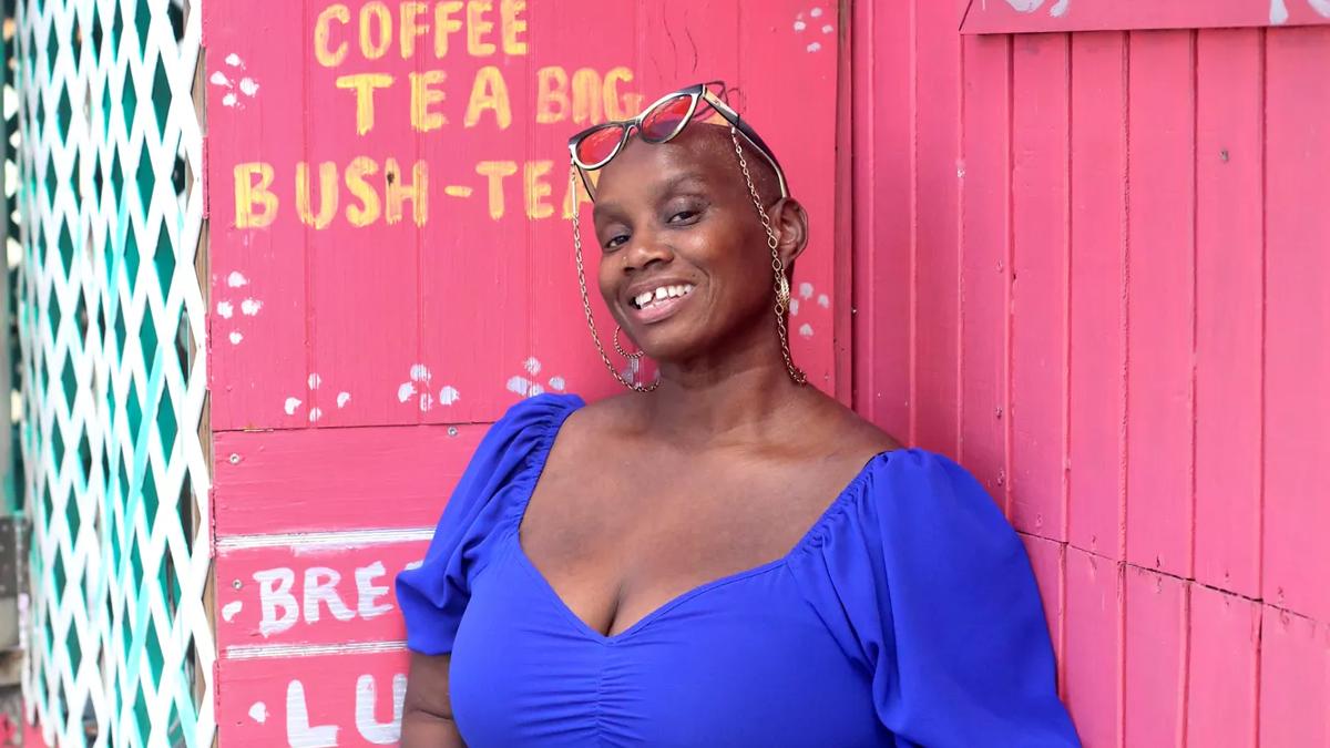 Andi Oliver leans against a pink food shack wearing a blue v-necked dress. She wears her glasses on her head and smiles to camera. 