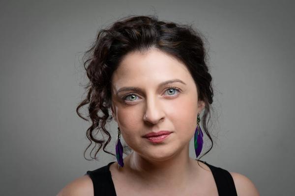 Portrait of author Christy Lefteri against a plain grey background. She has her hair tied back with some loose curls at the front. and is wearing purple feathers as earrings.