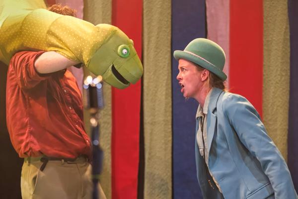 Women in a blue suit and bowler hat shouts at a dinosaur puppet.