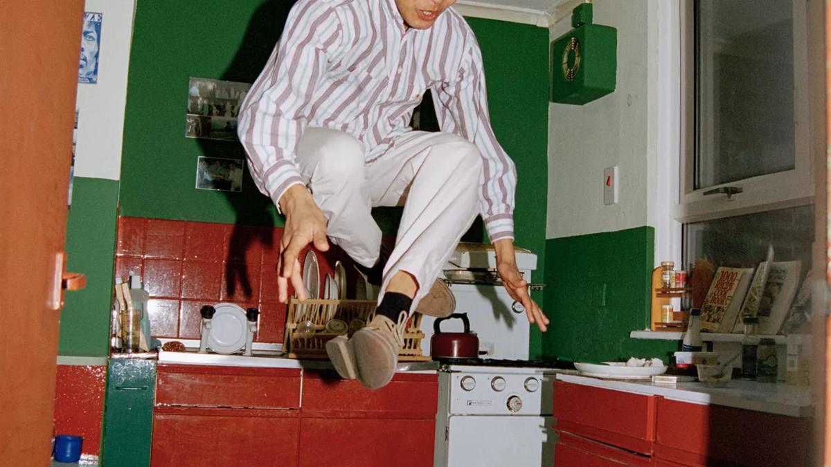 A man leaps into the air in a crouched position in a domestic kitchen that has a red and green floor, red units and a green wall