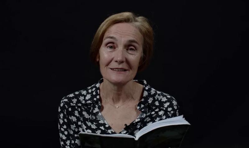 Jane Clarke, an older White woman with short brown hair and wearing a multi-coloured blouse, reads from their TS Eliot Prize shortlisted collection against a black background