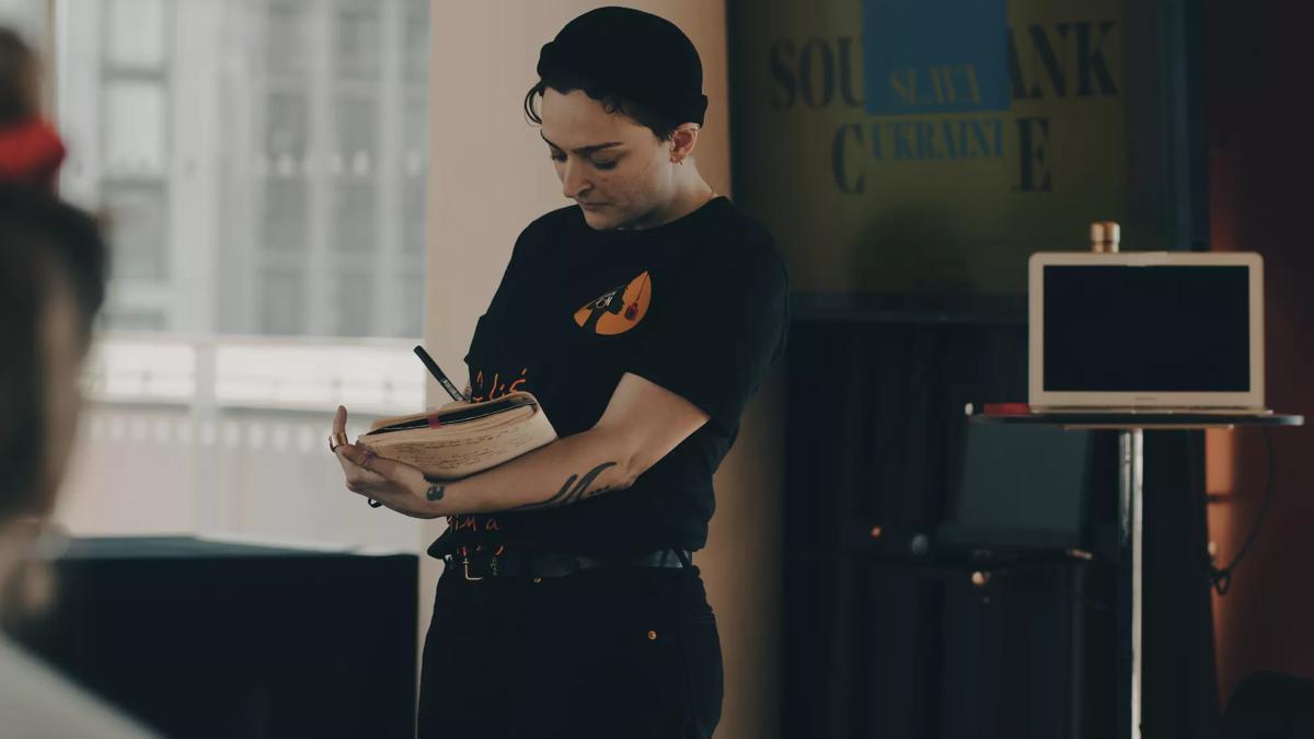 A woman with tattooed arms wearing black jeans, a black t -shirt and a black beanie stands up and writes in an A4 notebook. Behind her a silver MacBook laptop is open on a stand and a TV screen with the Southbank Centre logo sits at the back of the image. 