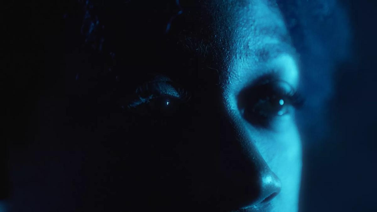 Tinted blue image of a close up of a Black woman's eyes. The left side of her face is shaded in darkness. 