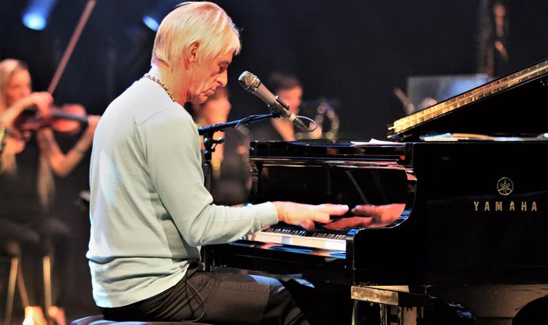 Paul Weller plays piano on the stage of the Royal Festival Hall, with an orchestra playing in the background