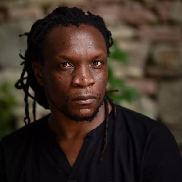 The poet Ishion Hutchinson, a young Black man with his hair back in in tight dreadlocks  wears a black shirt in front of a brick wall