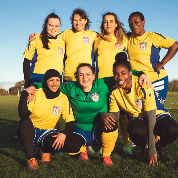 A diverse group of 7 women in yellow and blue football kits pose for a team photo in a field. 