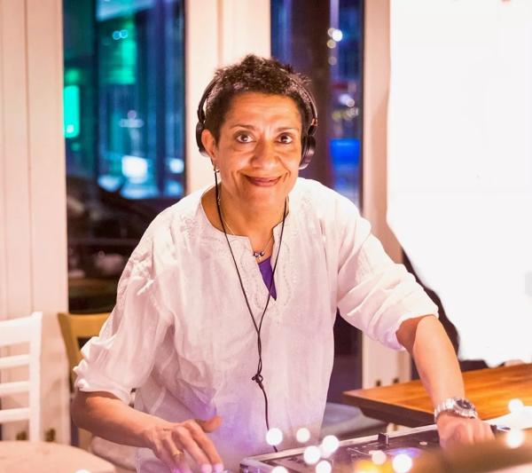 DJ Ritu stands at the decks with headphones on in a white shirt smiling at the camera. 