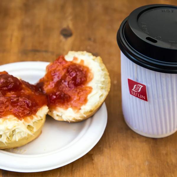 Photo of a scone covered in clotted cream on the bottom and jam on top, next to a takeaway cup of tea, served by Tea, Bread & Brownie at Southbank Centre Food Market
