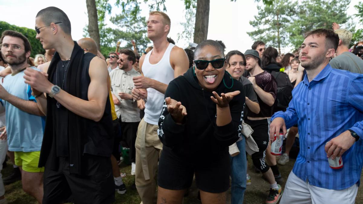 People dancing outdoors at a London Queer Sound System event. Person wearing black sunglasses and black shorts and jumper looking at the camera with their tongue out.